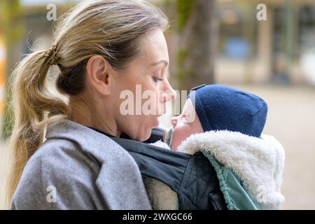 SideView einer attraktiven blonden Spätmutter in ihren 40ern, die ihr neugeborenes Baby liebevoll in einer Babytrage hält Stockfoto