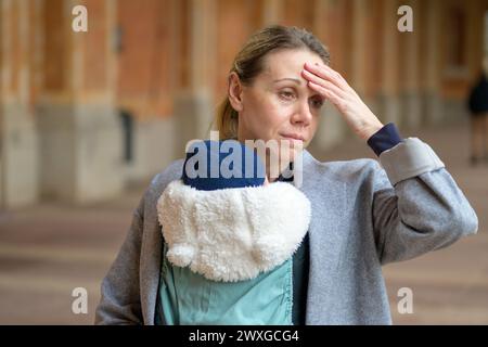 Erschöpfte und gestresste spätgeborene Mutter in ihren 40ern hält ihr neugeborenes Baby in einer Babytrage und sieht im Freien genervt aus Stockfoto