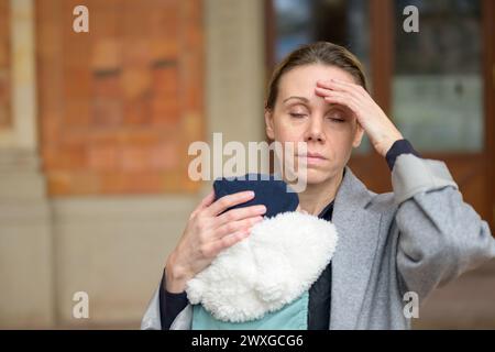 Erschöpfte und gestresste spätgeborene Mutter in ihren 40ern hielt ihr neugeborenes Baby in einer Babytrage und sah mit geschlossenen Augen genervt auf die Kamera Outdo aus Stockfoto