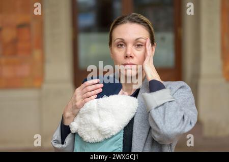 Erschöpfte und gestresste spätgeborene Mutter in ihren 40ern, die ihr neugeborenes Baby in einer Babytrage hält und verärgert auf die Kamera im Freien blickt Stockfoto