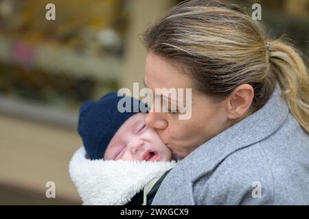 Nahaufnahme einer glücklichen Mutter in ihren 40ern, die ihr weinendes neugeborenes Baby in einer Babytrage hält und es liebevoll im Freien küsst Stockfoto