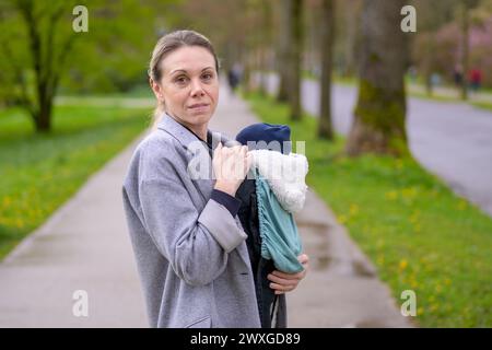 Erschöpfte und gestresste spätgeborene Mutter in ihren 40ern, die ihr neugeborenes Baby in einer Babytrage hält und in einem Park genervt auf die Kamera blickt Stockfoto