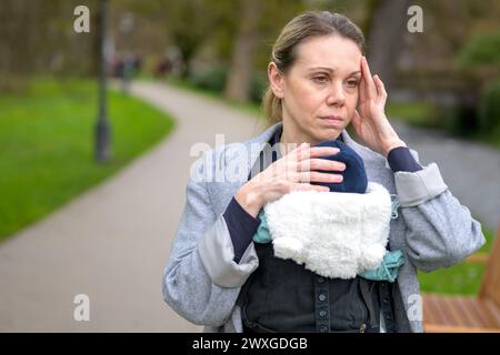 Erschöpfte und gestresste Frau, die ihr Baby in einer Babytrage im Park hält und trägt Stockfoto