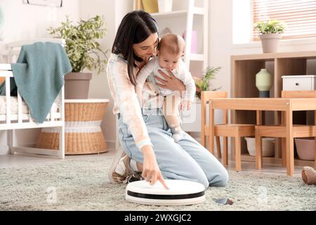 Junge Frau mit ihrem kleinen Baby und Roboter-Staubsauger auf Teppich im Schlafzimmer Stockfoto