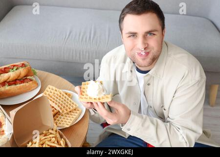 Junger Mann in Sahne, der zu Hause Waffeln isst. Das Konzept des Überessens Stockfoto