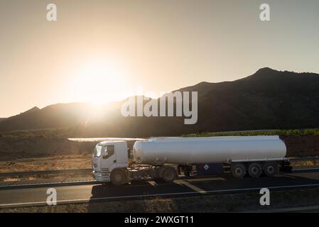 Weißer Tankwagen, der auf einer Autobahn fährt, während die Sonne zwischen den Bergen aufgeht. Stockfoto