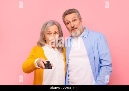 Schockiertes Seniorenpaar, Das Fernsehen Mit Fernbedienung Sieht, Pink Studio Stockfoto