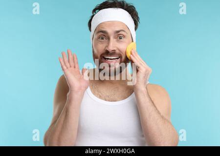Mann mit Stirnband, der sein Gesicht mit Schwamm auf hellblauem Hintergrund wäscht Stockfoto