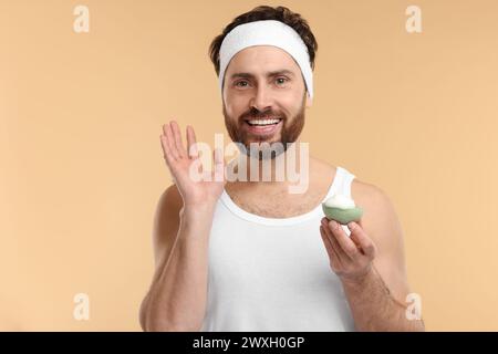 Mann mit Stirnband, der sein Gesicht mit einem Schwamm auf beigem Hintergrund wäscht Stockfoto