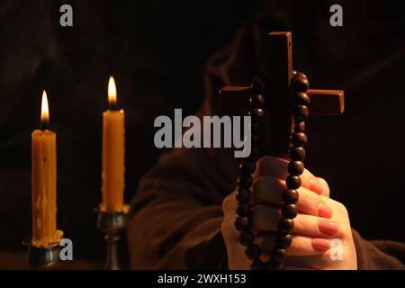 Frau mit Kreuz, Perlen und brennenden Kerzen betend auf verschwommenem Hintergrund, Nahaufnahme Stockfoto