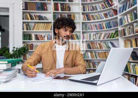 Ernsthafter junger Mann mit lockigen Haaren, der sich darauf konzentriert, mit einem Laptop in einer Bibliothek zu lernen, mit Büchern um ihn herum. Stockfoto