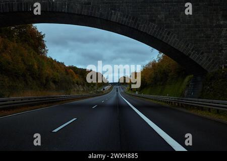 Der Bogen der Brücke über die Straße erstreckt sich bis in die Ferne. Bäume und Sträucher mit Herbstlaub am Straßenrand. Deutschland. Stockfoto