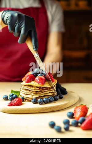 Mann, der Creme Pancakes auf einem Holzbrett mit Erdbeeren und Heidelbeeren schneidet, warmes Licht, Bar Stockfoto
