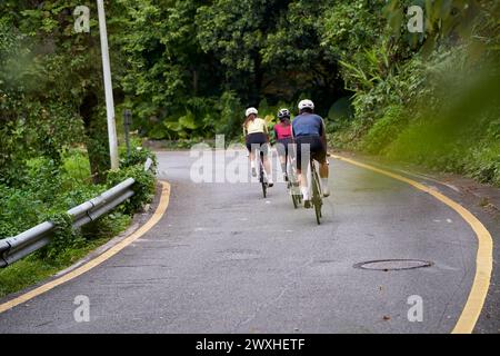 Rückansicht einer Gruppe von drei jungen asiatischen erwachsenen Radfahrern, die im Freien auf der Landstraße Fahrrad fahren Stockfoto