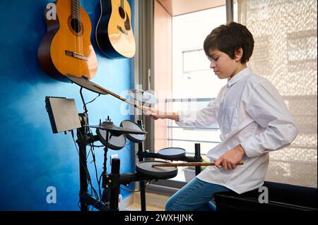 Teenager in weißem Hemd und blauer Jeans, der Schlagzeug in seinem modernen Musikstudio spielt. Zwei akustische elektrische Gitarren hängen an einer blauen Wand. Stockfoto