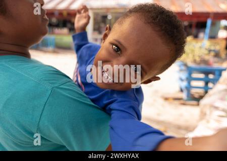 INDONESIEN, INSEL KRI - 27. JANUAR 2024: Ein kleiner Papua-Junge mit funkelnden Augen sitzt in seiner Mutter Stockfoto