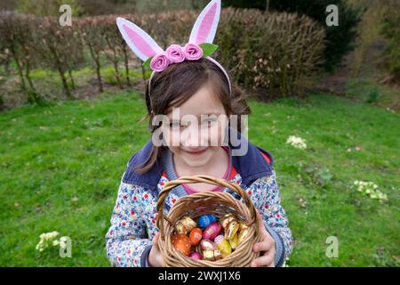 Ostereierjagd lächelndes Mädchen 6 7 mit Kaninchenohren, die einen Korb mit Schokoladeneiern halten, in Wales March Garden 2024 Großbritannien KATHY DEWITT Stockfoto