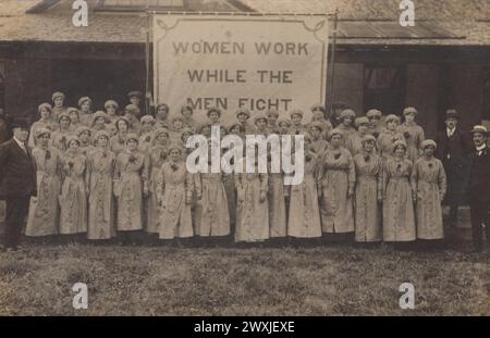 „Women work while the Men Fight“: Foto von Munitionsarbeiterinnen außerhalb der Siemens-Fabrik in Stafford während des Ersten Weltkriegs (wahrscheinlich 1916 oder 1917). Die Frauen und das Banner, vor dem sie stehen, nahmen an dem jährlichen Krankenhauswettbewerb oder -Parade Teil. Die Munitionsarbeiterinnen tragen alle ihre Arbeitskleidung - lange Overalls und eng anliegende Kappen - und haben einen Zweig Blumen oder Laub am Revers Stockfoto