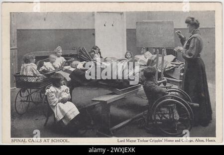 Lord Mayor Treloar Cripples' Home and College, Alton, Hampshire: Spinal Cases at School. Postkarte einer Gruppe von Kindern aus dem frühen 20. Jahrhundert in einem Klassenzimmer mit einer aufstehenden Frau, die die Klasse unterrichtet. Die Kinder befinden sich in einer Reihe von Positionen und verwenden verschiedene Arten von Spezialausrüstung, einschließlich Rollstuhl und Kinderrückenwagen Stockfoto