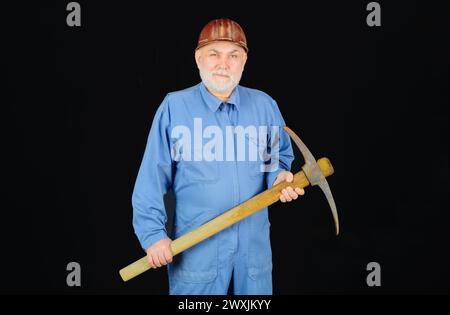 Bau- und Bauarbeiten. Bärtiger Mann in Overalls und Schutzhelm mit Pickel-Axt. Handwerker oder Bauunternehmer mit Spitzhacke. Männlicher Baumeister in Uniform Stockfoto