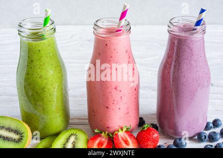 Sortiment an frischen Frucht-Smoothies oder Milchshake in Glasflaschen mit frischen Beeren und Stroh auf weißem Holzhintergrund. Rote, grüne und blaue Flaschen Stockfoto