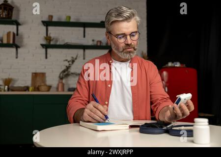 Diabetiker, der den Blutzuckerspiegel mit einem Glukometer überprüft, macht Notizen mit Indikatoren im Notizblock Stockfoto