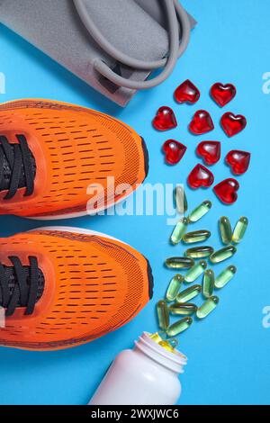 Gelbe Kapseln mit ungesättigten Fettsäuren und roten Glasherzen, Tonometer zur Blutdruckmessung, sportliche orangefarbene Sneakers auf blauem Hintergrund Stockfoto