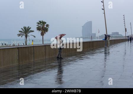 Barcelona, Spanien. 31. März 2024. Eine Frau geht durch Barceloneta und schützt sich während des Sturms Nelson vor dem Regen mit einem Regenschirm. Blick auf den Strand Barceloneta in Barcelona, Spanien während des Sturms Nelson, der zehn spanische Gemeinden während dieser Karwoche wegen Regen, Wind oder Schnee auf gelber Alarmstufe gehalten hat. Quelle: SOPA Images Limited/Alamy Live News Stockfoto