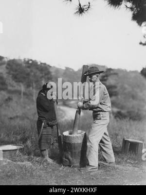 In Camp John Hay, gebürtiger Barrio, Baguio, Philippinen, wurde eine Ifugao-Frau aufgenommen, die einem amerikanischen Soldaten dabei zusieht, wie er sich beim Stampfen von Reis Ca versucht. 1931 Stockfoto