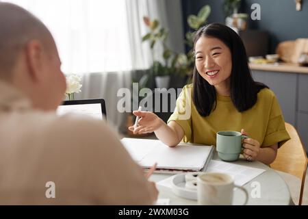 Über-Schulter-Aufnahme einer fröhlichen jungen asiatischen Frau, die mit ihrem Freund plaudert, während sie gemeinsam Hausaufgaben macht Stockfoto