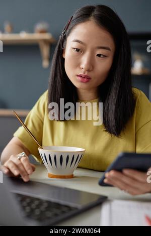 Vertikales Porträt einer jungen asiatischen Freelancerin, die während des Frühstücks ein Smartphone hält, das an einem Laptop arbeitet Stockfoto