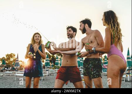 Fröhliche Freunde, die mit Bierdosen am Strand planschen, verkörpern den Geist des Sommerspaßes, während der Tag in einen wunderschönen Sonnenuntergang abgeht - Junge Leute sozia Stockfoto