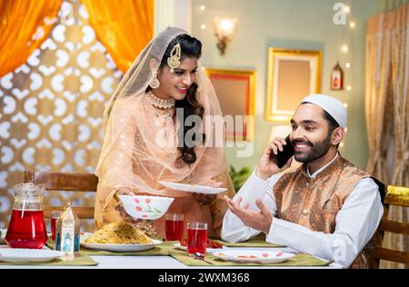 Glücklicher indischer Ehemann, der Freunde oder Verwandte anruft, während seine Frau Ramadan-Essen oder Festmahl bei Festivalfeiern organisiert - Konzept der Technologie Stockfoto