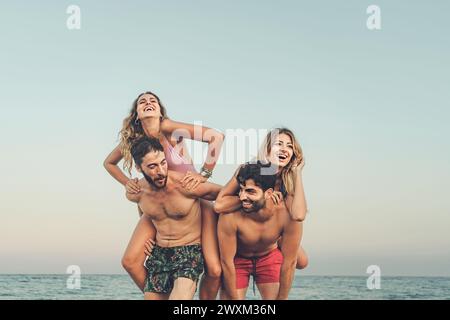 Zwei Paare lachen mit Huckepack-Fahrten am Strand und verkörpern den Geist eines unbeschwerten und fröhlichen Urlaubs am Meer - Kopierraum in der SK Stockfoto