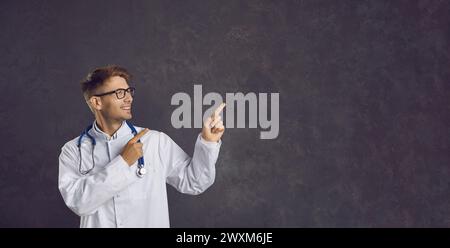 Ein glücklicher junger Doktor im weißen Mantel, der seine Finger auf den Hintergrund des Kopierraums zeigt Stockfoto