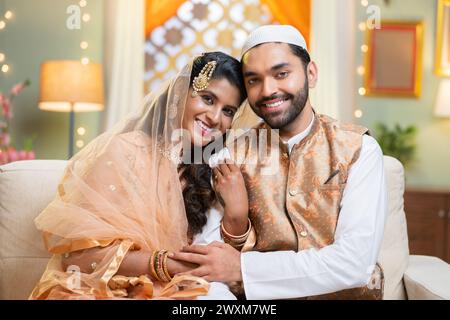 Happy smiling indain Muslim couple embracing by looking at camera while sitting on sofa at home - concept of traditional culture, Ramadan greeting and Stockfoto