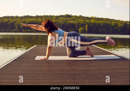 Eine übergewichtige Frau macht Plank-Bewegung, während sie Yoga auf dem See in der Natur übt Stockfoto