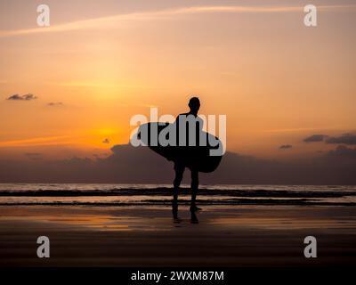 Mann-Silhouette mit Surfbrett, Spaziergang an der dunklen Küste. Surfer steht am Sonnenuntergang über dem Meerwasserhintergrund. Sommer Sandstrand in Even Stockfoto