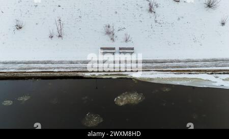 Drohnenfotografie von Bänken am Flussufer, die während des bewölkten Wintertages von Schnee bedeckt sind Stockfoto