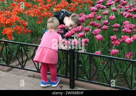 Moskau, Russland - 12. Mai 2018: Eine moderne Mutter spaziert mit ihrem Kind in einem öffentlichen Park. Mutter und Kind schauen mit Interesse auf Blumen in einem Blumenbeet Stockfoto