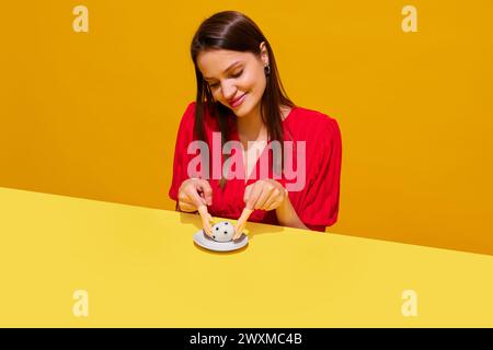 Junge lächelnde Frau, die kleine Spielzeughände hält und Ei mit google-Augen vor orange gelbem Hintergrund fängt. Spaß Stockfoto