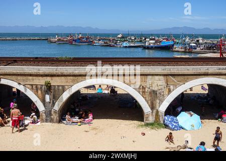 Südafrikaner genießen einen sonnigen Tag am Strand unter einer historischen Eisenbahnbrücke in Kalk Bay mit einem malerischen Hafen im Hintergrund. Stockfoto