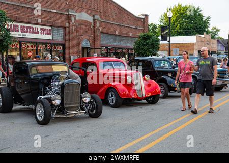 Ein paar spazieren vorbei an einem roten 1936 Chevrolet und einem schwarzen 1932 Ford Hotrod, der vor dem Country Lane General Store auf einer Autoausstellung in Auburn, Ind. Ausgestellt wird Stockfoto