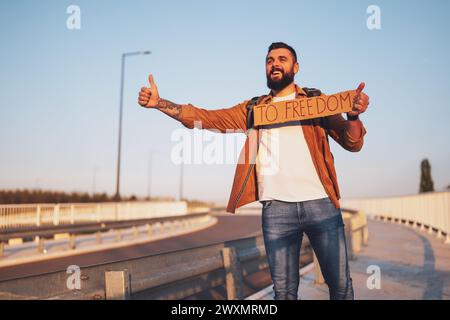 Der Mann trampt am Straßenrand und versucht, das Auto anzuhalten. Er hält Pappe mit Inschrift in der Hand. Stockfoto