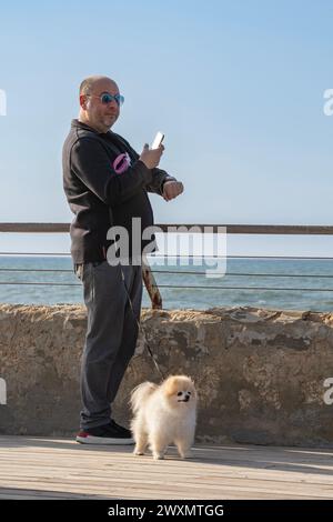 Tel Aviv, Israel - 6. Januar 2023: Ein Mann und sein pommerscher Hund auf dem Deck mit Blick auf das mittelmeer im Hafen von Tel Aviv an einem sonnigen Tag. Stockfoto