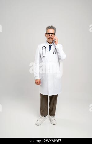 Der grauhaarige Arzt steht aufrecht, während er am Telefon spricht, und spiegelt die aktive Rolle der Ärzte bei der Koordinierung der Patientenversorgung auch aus der Entfernung wider. Stockfoto