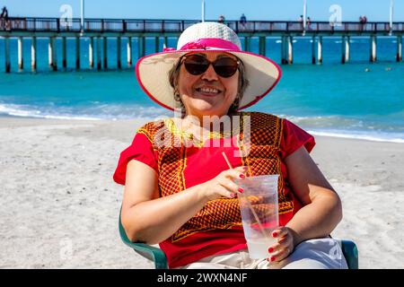 Lächelnde mexikanische Frau, die im Glas Kokoswasser trinkt, Pier und Ozean im Hintergrund, Hut, Sonnenbrille und rote Bluse, Su genießt Stockfoto