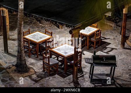 Blick aus der Vogelperspektive auf die quadratischen Tische und Holzstühle auf der Terrasse des kleinen Restaurants, beleuchtet durch Lampen, Steinboden, Metallgrill, ruhige Nacht im Hotel Stockfoto