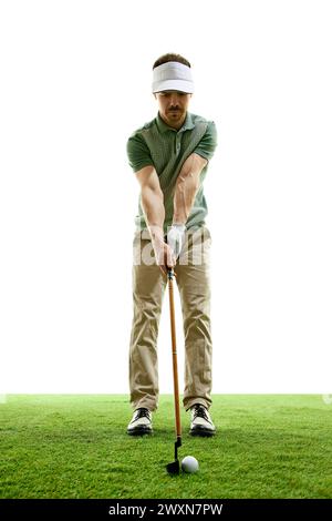 Fokussierter Golfer zu Beginn der kraftvollen Fahrt vor weißem Studiohintergrund. Ein erfahrener Golfspieler steht mit einem Golfschläger auf grünem Gras. Stockfoto
