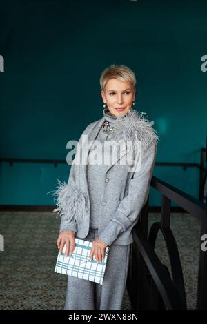 Stilvolle, reife Frau mit kurzem Haarschnitt, gekleidet in grauem Strickanzug mit Jacke, Federn und modischen Accessoires, Clutch halten, gegen Emera Stockfoto